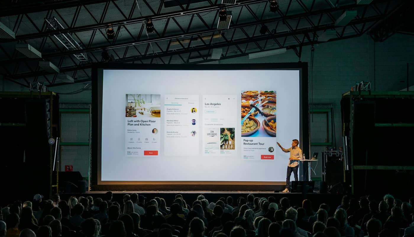 A speaker presenting on a stage in front of a large projected screen and an audience